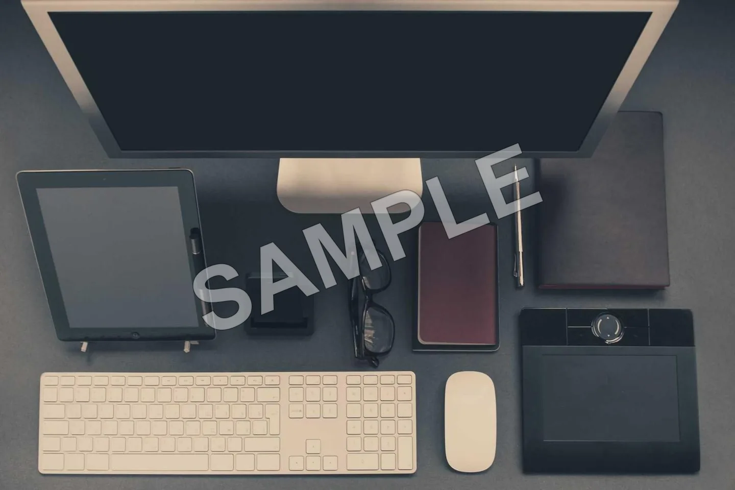 Overhead photograph of a desk with a monitor, tablet and keyboard, crossed by a diagonal SAMPLE banner